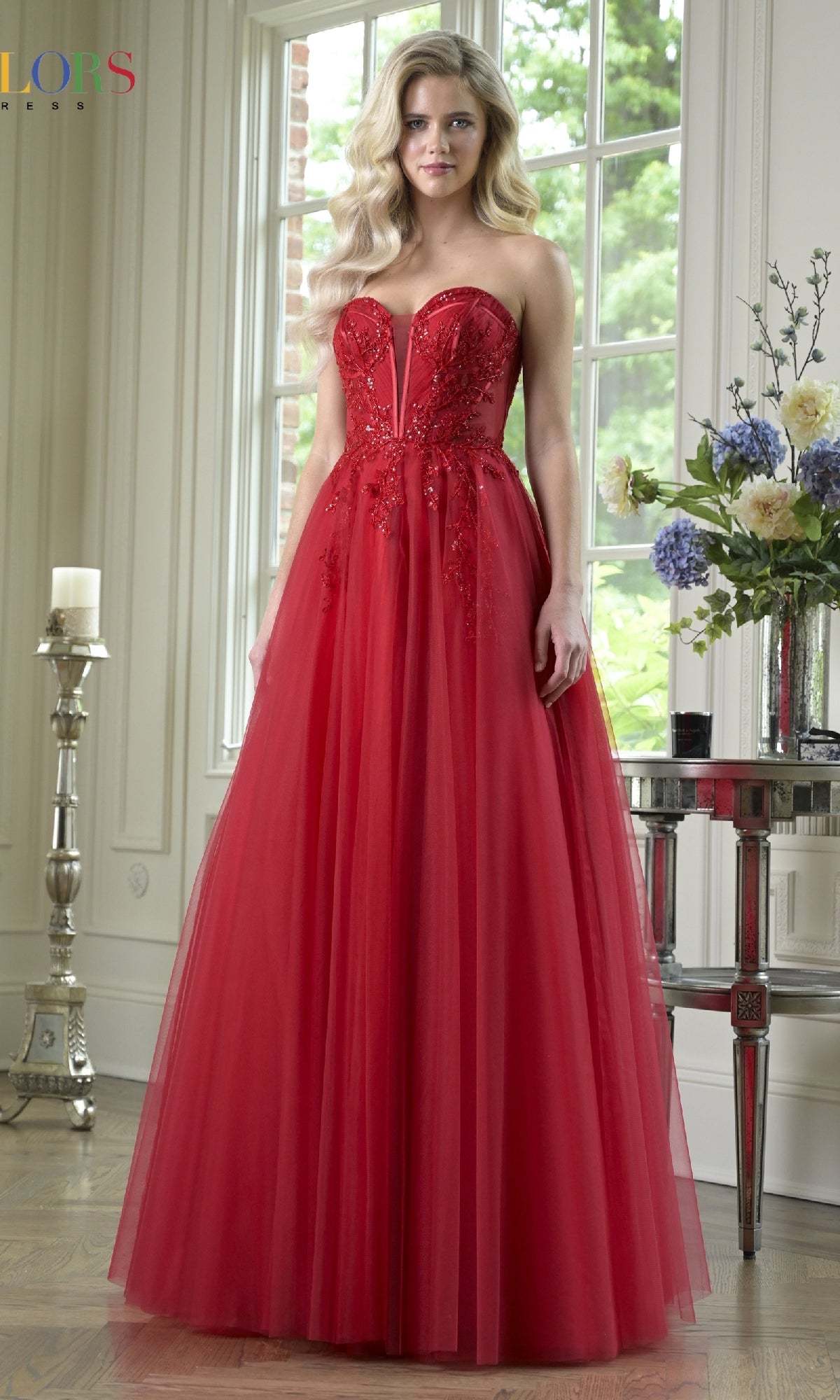 A woman stands indoors wearing the Strapless Long Corset Prom Dress: Colors Dress 3923, featuring a fitted bodice and tulle skirt in red; flowers and a window appear in the background.